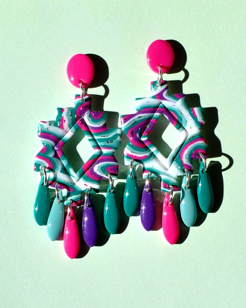 Paire de boucles d'oreilles iconique présentée sur Instagram, en trois partie: une puce rose vif uni qui surmonte une forme géométrique "précolombienne" motif "à la dérive des astres" surmontant cinq pampilles unie (vert vague marine, rose unie, violet et deux pampilles vert émeraude)