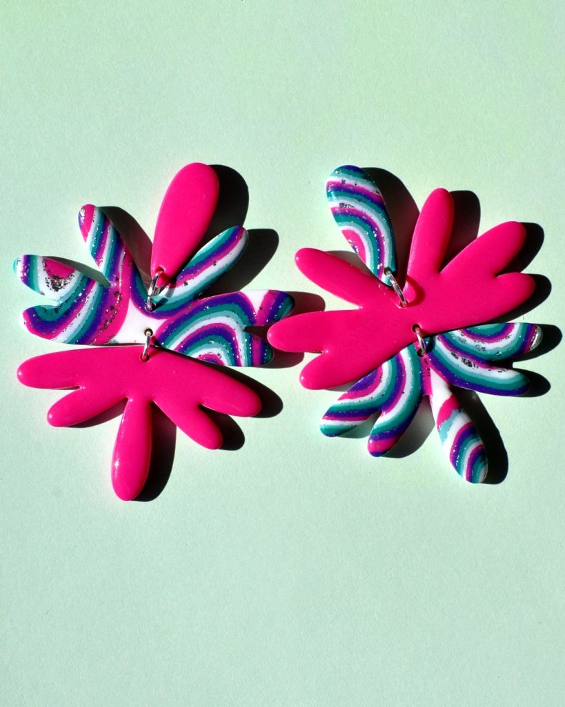 Paire de boucles d'oreilles en forme de "fleur maximaliste" abstraite asymétrique en trois parties. La puce forme un pétale rose uni vif. Le corps de la boucle est composé de cinq pétales au motif "à la dérive des astres", surmontant cinq autres pétales rose unie. L'autre boucle est en inversé (puce pétale d'accroche motif "à la dérive des astres", corps de cinq pétales rose uni et énorme pampille de cinq pétales au motif "à la dérive des astres").