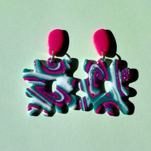 Paire de boucles d'oreilles en forme de "carré thwomp" aux bords crantés, ajourée au milieu, au motif "à la dérive des astres", surmontée d'une puce en longueur de forme abstraite rose vif.