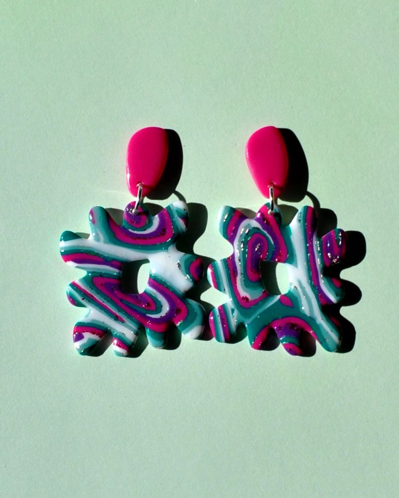 Paire de boucles d'oreilles en forme de "carré thwomp" aux bords crantés, ajourée au milieu, au motif "à la dérive des astres", surmontée d'une puce en longueur de forme abstraite rose vif.