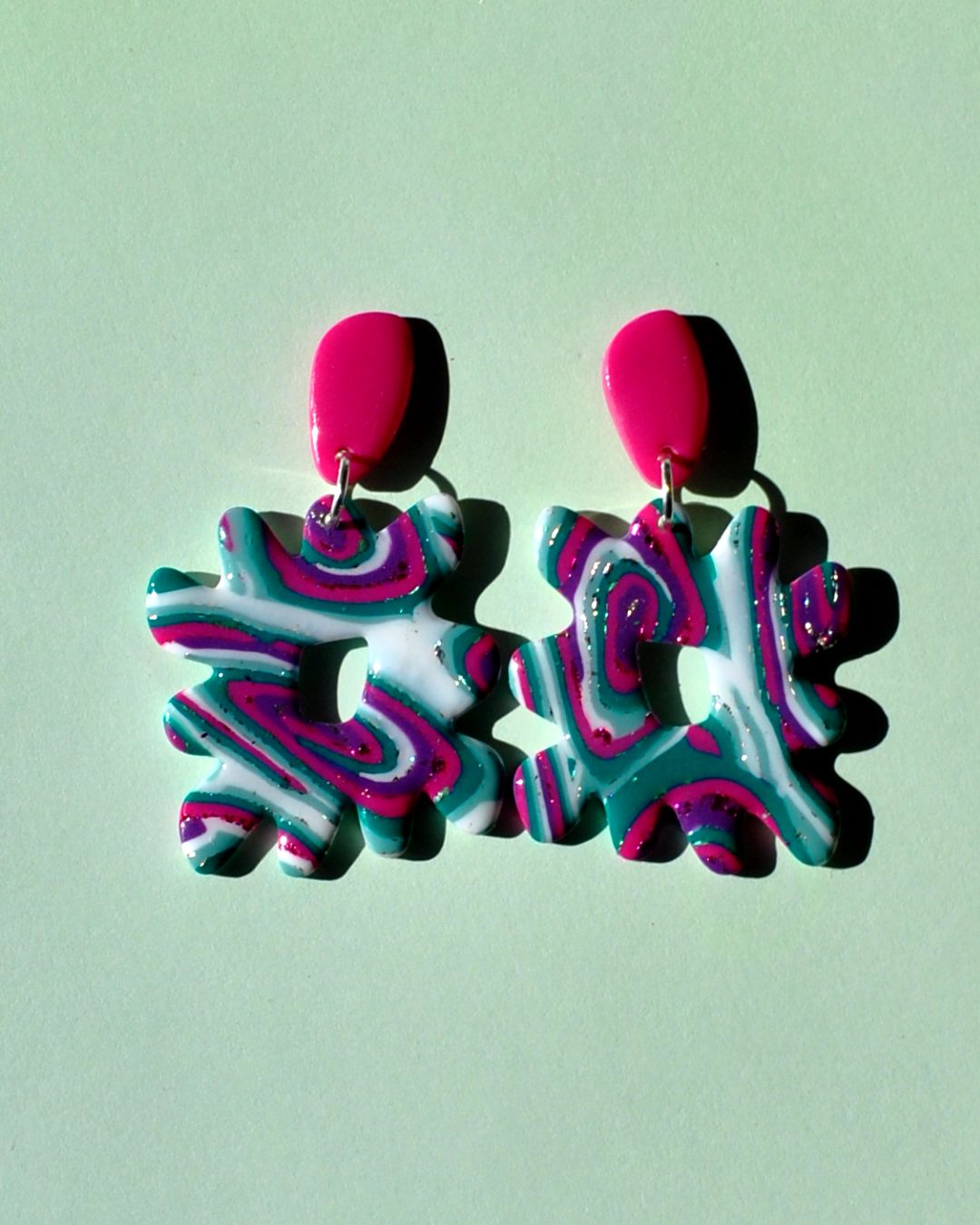 Paire de boucles d'oreilles en forme de "carré thwomp" aux bords crantés, ajourée au milieu, au motif "à la dérive des astres", surmontée d'une puce en longueur de forme abstraite rose vif.
