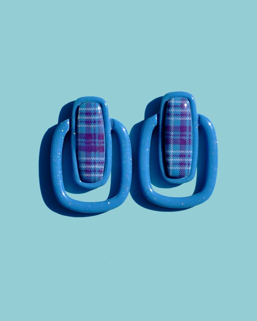 Paire de boucles d'oreilles en forme de rectangle long, cerclé d'un autre rectangle plus bas, plus fin arrondi. Il y a un jour entre les deux rectangles. La paire de boucles d'oreilles est unie, d'un bleu glacé. Le premier rectangle possède un rectangle en relief "cabochon" au motif à carreaux "check" lie-de-vin et blancs.