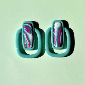 Paire de boucles d'oreilles en forme de rectangle long, cerclé d'un autre rectangle plus bas, plus fin arrondi. Il y a un jour entre les deux rectangles. La paire de boucles d'oreilles est unie vert vague marine. Le premier rectangle possède un rectangle en relief au motif "à la dérive des astres"