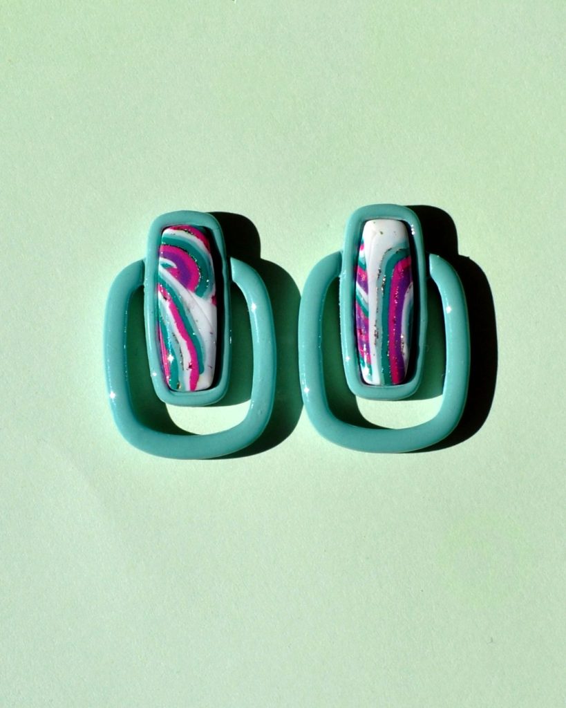 Paire de boucles d'oreilles en forme de rectangle long, cerclé d'un autre rectangle plus bas, plus fin arrondi. Il y a un jour entre les deux rectangles. La paire de boucles d'oreilles est unie vert vague marine. Le premier rectangle possède un rectangle en relief au motif "à la dérive des astres"