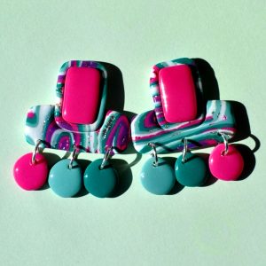 Paire de boucles d'oreilles en forme double rectangle imbriqué (un en longueur, l'autre en largeur). La paire de boucles d'oreilles est au motif "à la dérive des astres". Le premier rectangle en longueur possède un rectangle en relief uni rose vif. Du rectangle en largeur partent trois petites pampilles rondes irrégulières rose vif, vert vague marine, vert émeraude.