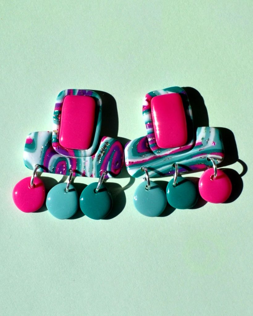 Paire de boucles d'oreilles en forme double rectangle imbriqué (un en longueur, l'autre en largeur). La paire de boucles d'oreilles est au motif "à la dérive des astres". Le premier rectangle en longueur possède un rectangle en relief uni rose vif. Du rectangle en largeur partent trois petites pampilles rondes irrégulières rose vif, vert vague marine, vert émeraude.