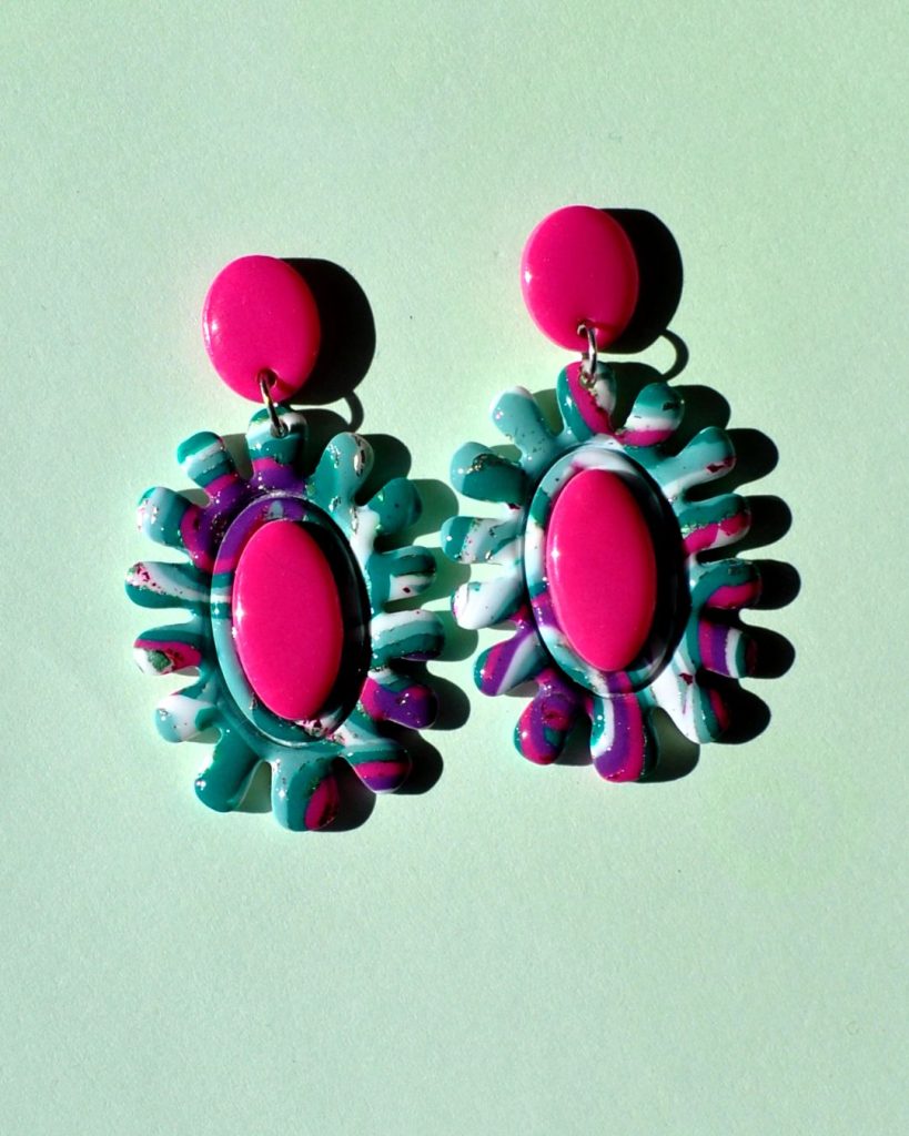 Paire de boucles d'oreilles en forme de fleur abstraite au motif "à la dérive des astres". Le cœur de la "fleur" est un ovale en longueur, en relief uni rose vif. La boucle est surmontée d'une puce ovale unie, rose vif.