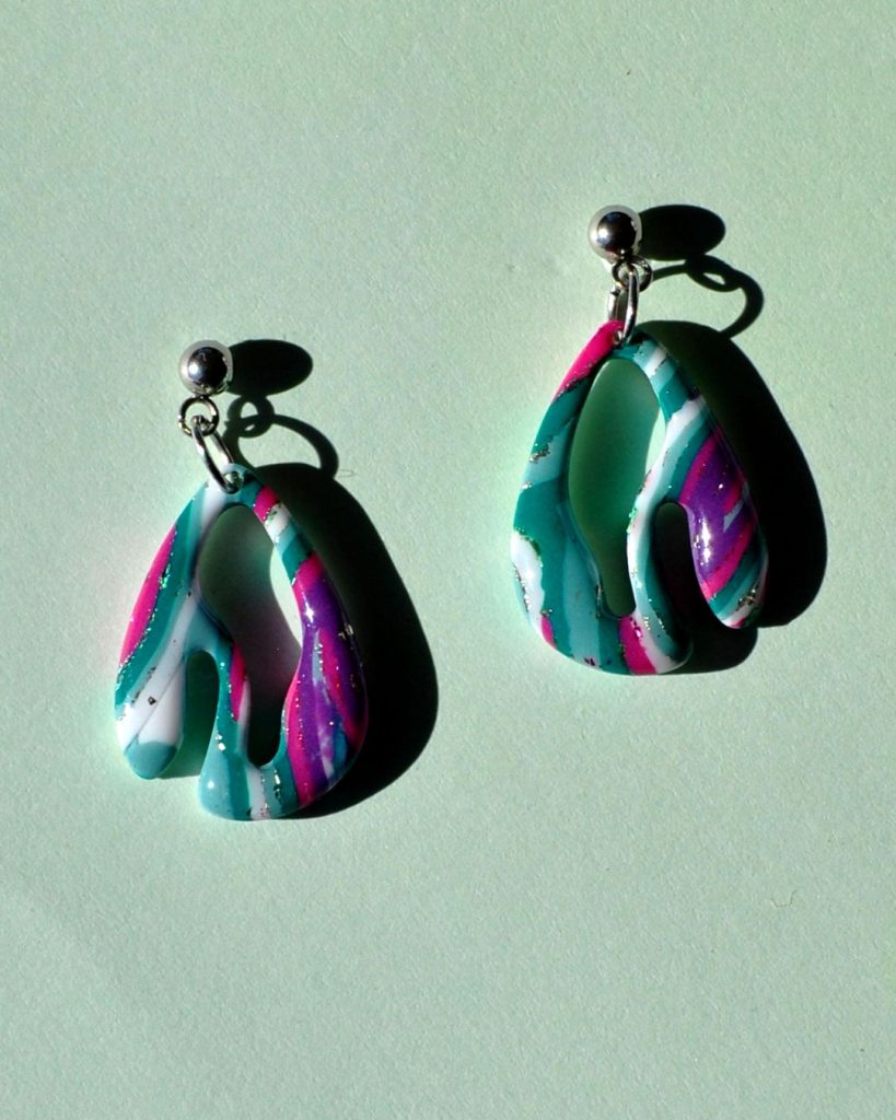 Petite paire de boucles d'oreilles en deux parties. Une petite puce en acier inoxydable surmonte une forme abstraite au motif "à la dérive des astres".