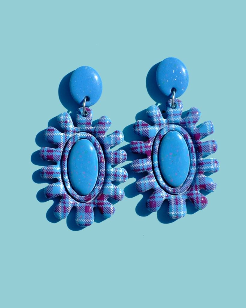 Paire de boucles d'oreilles en forme de fleur abstraite au motif carreaux "check" blancs et lie-de-vin. Le cœur de la "fleur" est un ovale en longueur, en relief uni bleu glacé. La boucle est surmontée d'une puce ovale unie, bleue glacée.