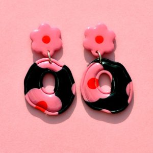 Paire de boucles d'oreilles ronde abstraite rétro avec un liseret incrusté. La paire de boucles d'oreilles est ajourée au centre. Une puce fleur rose à cœur rouge vif surmonte une boucle à fond noir avec les mêmes fleurs roses à cœur rouge.