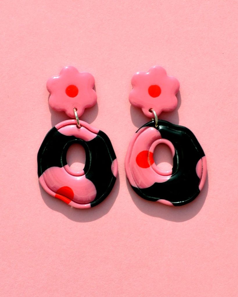 Paire de boucles d'oreilles ronde abstraite rétro avec un liseret incrusté. La paire de boucles d'oreilles est ajourée au centre. Une puce fleur rose à cœur rouge vif surmonte une boucle à fond noir avec les mêmes fleurs roses à cœur rouge.