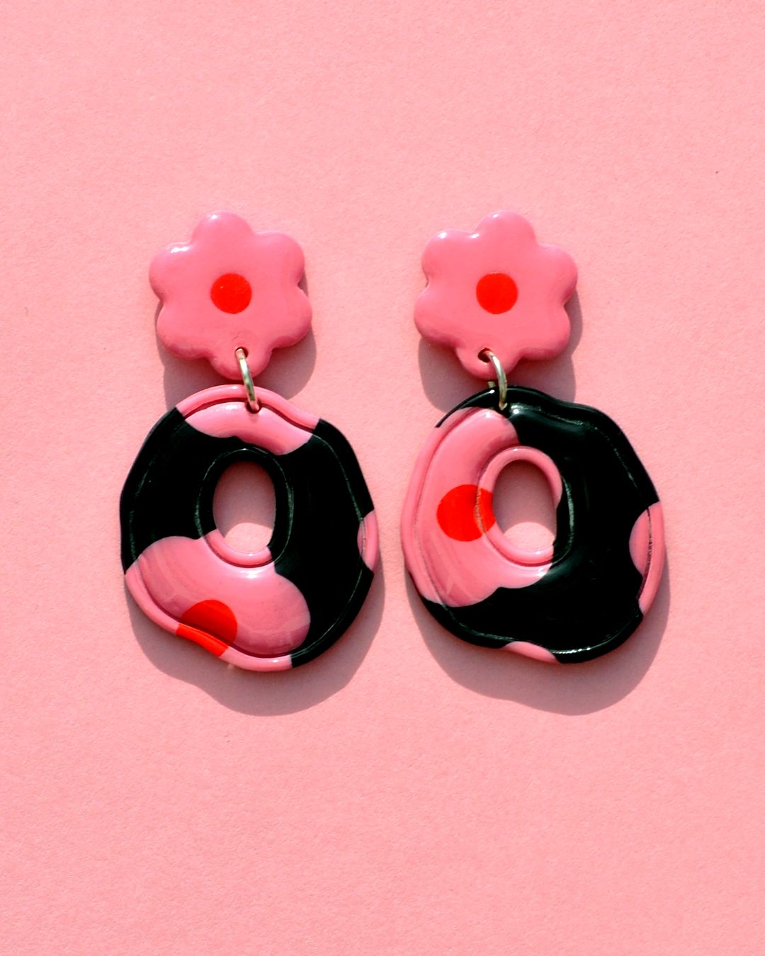 Paire de boucles d'oreilles ronde abstraite rétro avec un liseret incrusté. La paire de boucles d'oreilles est ajourée au centre. Une puce fleur rose à cœur rouge vif surmonte une boucle à fond noir avec les mêmes fleurs roses à cœur rouge.