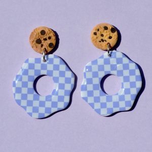 Paire de boucles d'oreilles en forme de fleur abstraite. La paire de boucles d'oreilles est ajourée au centre d'un cercle bien défini. Une puce cookie surmonte une boucle motif damier violet pâle sur fond blanc.