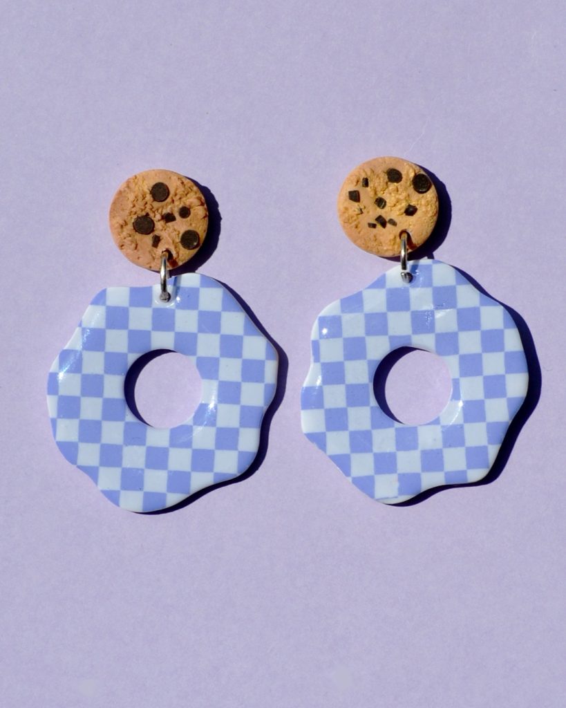 Paire de boucles d'oreilles en forme de fleur abstraite. La paire de boucles d'oreilles est ajourée au centre d'un cercle bien défini. Une puce cookie surmonte une boucle motif damier violet pâle sur fond blanc.