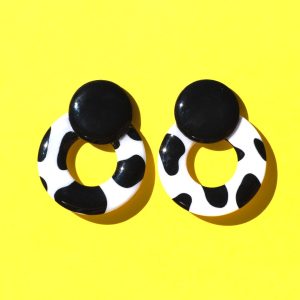 Paire de boucles d'oreilles en forme de cercle ajouré. Une grosse puce ronde en relief est collée sur le haut de la boucle. La boucle est au motif tâches de vache noires sur fond blanc. La "puce" ronde est noire unie.