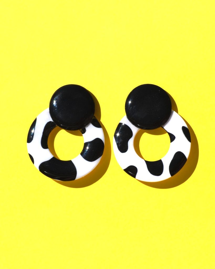 Paire de boucles d'oreilles en forme de cercle ajouré. Une grosse puce ronde en relief est collée sur le haut de la boucle. La boucle est au motif tâches de vache noires sur fond blanc. La "puce" ronde est noire unie.