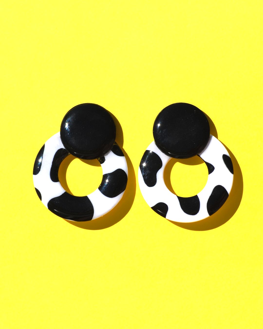 Paire de boucles d'oreilles en forme de cercle ajouré. Une grosse puce ronde en relief est collée sur le haut de la boucle. La boucle est au motif tâches de vache noires sur fond blanc. La "puce" ronde est noire unie.