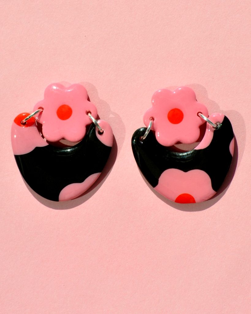 Paire de boucles d'oreilles en forme de courbe épaisse. Une puce fleur rose à cœur rouge vif surmonte une boucle à fond noir avec les mêmes fleurs roses à cœur rouge.