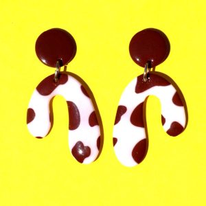 Paire de boucles d'oreilles en forme abstraite motif tâches de vache brunes sur fond blanc, surmontée d'une puce ronde marron.