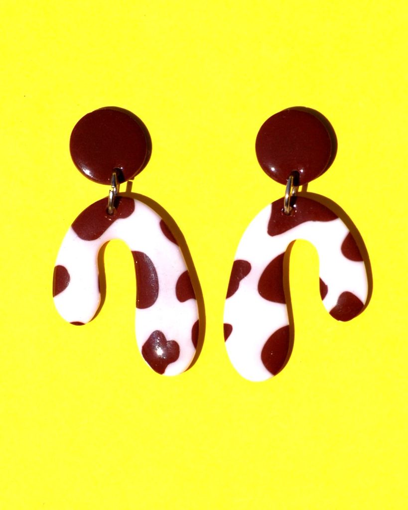 Paire de boucles d'oreilles en forme abstraite motif tâches de vache brunes sur fond blanc, surmontée d'une puce ronde marron.