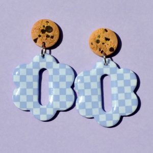 Paire de boucles d'oreilles en forme de grosse fleur à six pétales symétriques. La paire de boucles d'oreilles est ajourée au centre d'un rectangle allongé arrondis sur les bords. Une puce cookie surmonte une boucle motif damier blanc sur fond violet pâle.