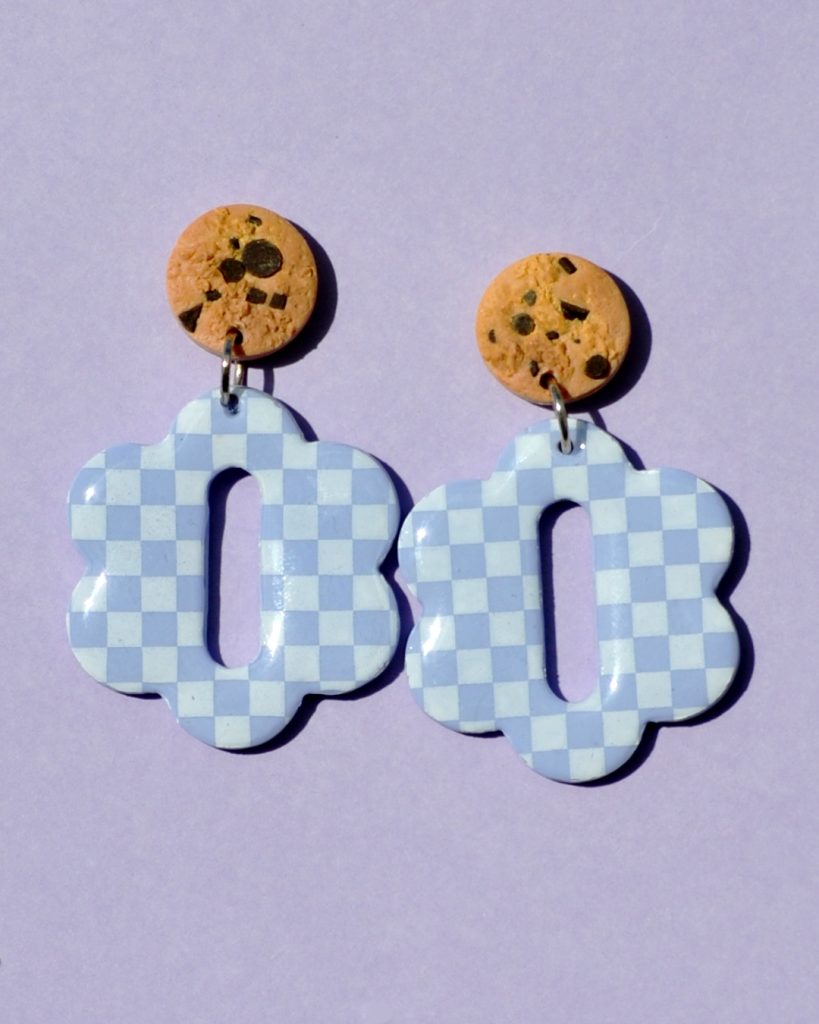 Paire de boucles d'oreilles en forme de grosse fleur à six pétales symétriques. La paire de boucles d'oreilles est ajourée au centre d'un rectangle allongé arrondis sur les bords. Une puce cookie surmonte une boucle motif damier blanc sur fond violet pâle.