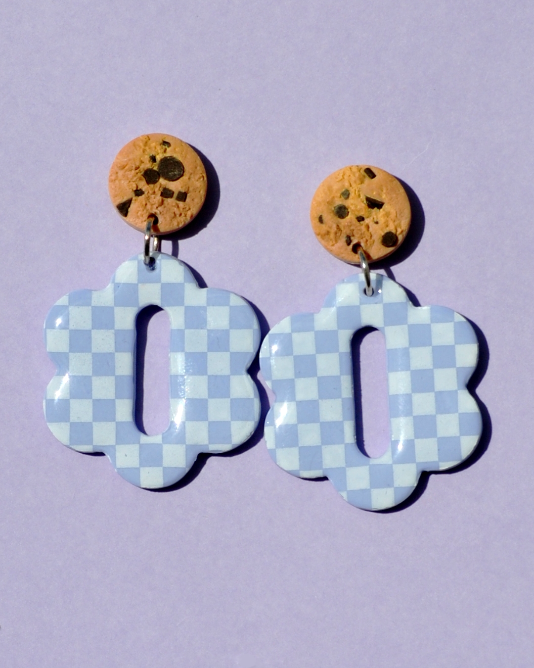 Paire de boucles d'oreilles en forme de grosse fleur à six pétales symétriques. La paire de boucles d'oreilles est ajourée au centre d'un rectangle allongé arrondis sur les bords. Une puce cookie surmonte une boucle motif damier blanc sur fond violet pâle.