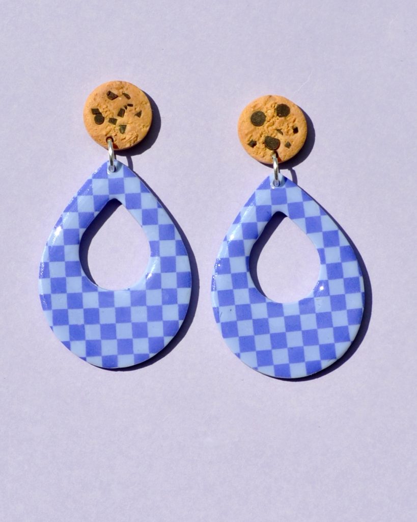 Paire de boucles d'oreilles d'énorme goutte dodue. La paire de boucles d'oreilles est ajourée au centre d'une goutte de taille moyenne. Une puce cookie surmonte une boucle motif damier violet soutenu sur fond violet pâle.