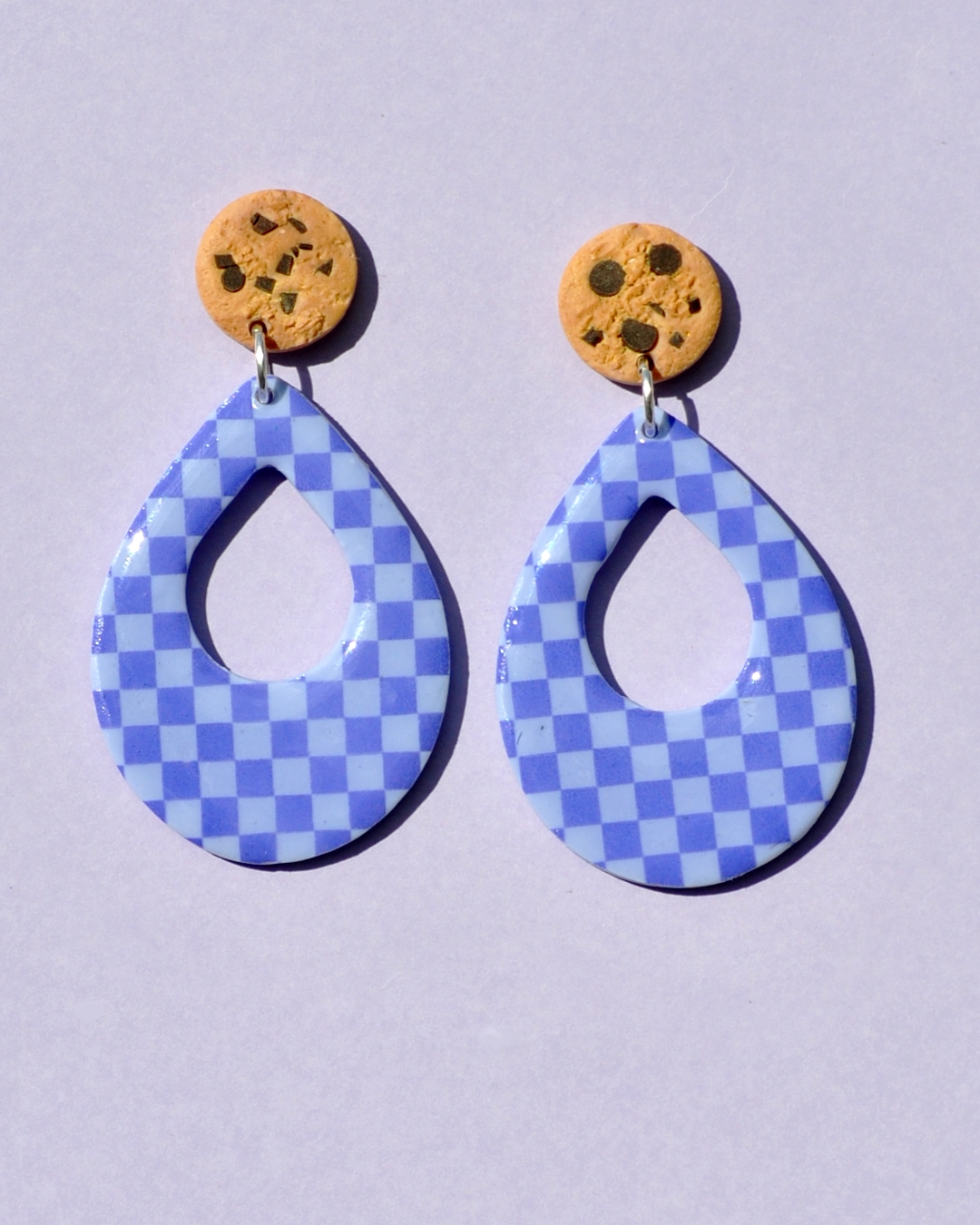 Paire de boucles d'oreilles d'énorme goutte dodue. La paire de boucles d'oreilles est ajourée au centre d'une goutte de taille moyenne. Une puce cookie surmonte une boucle motif damier violet soutenu sur fond violet pâle.