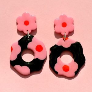 Paire de boucles d'oreilles en forme de fleur abstraite. La paire de boucles d'oreilles est ajourée au centre d'un cercle bien défini. Une puce fleur rose à cœur rouge vif surmonte une boucle à fond noir avec les mêmes fleurs roses à cœur rouge.