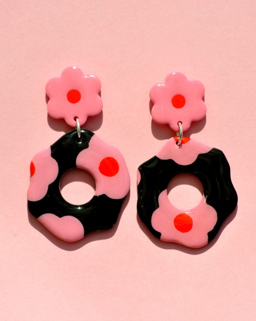 Paire de boucles d'oreilles en forme de fleur abstraite. La paire de boucles d'oreilles est ajourée au centre d'un cercle bien défini. Une puce fleur rose à cœur rouge vif surmonte une boucle à fond noir avec les mêmes fleurs roses à cœur rouge.