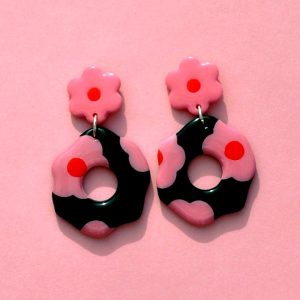 Paire de boucles d'oreilles en forme de fleur abstraite. La paire de boucles d'oreilles est ajourée au centre d'un cercle bien défini. Une puce fleur rose à cœur rouge vif surmonte une boucle à fond noir avec les mêmes fleurs roses à cœur rouge.