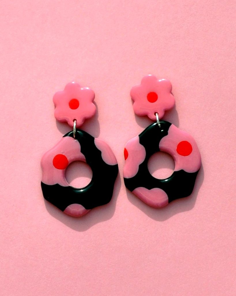 Paire de boucles d'oreilles en forme de fleur abstraite. La paire de boucles d'oreilles est ajourée au centre d'un cercle bien défini. Une puce fleur rose à cœur rouge vif surmonte une boucle à fond noir avec les mêmes fleurs roses à cœur rouge.