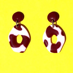 Paire de boucles d'oreilles en forme petit galet donut. La paire de boucles d'oreilles est ajourée au centre d'un cercle abstrait. Une petite puce ronde cookie surmonte une boucle motif tâches de vache brunes sur fond blanc.