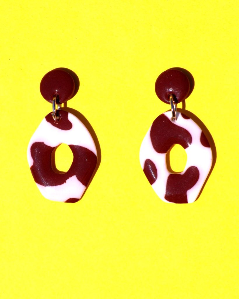 Paire de boucles d'oreilles en forme petit galet donut. La paire de boucles d'oreilles est ajourée au centre d'un cercle abstrait. Une petite puce ronde cookie surmonte une boucle motif tâches de vache brunes sur fond blanc.
