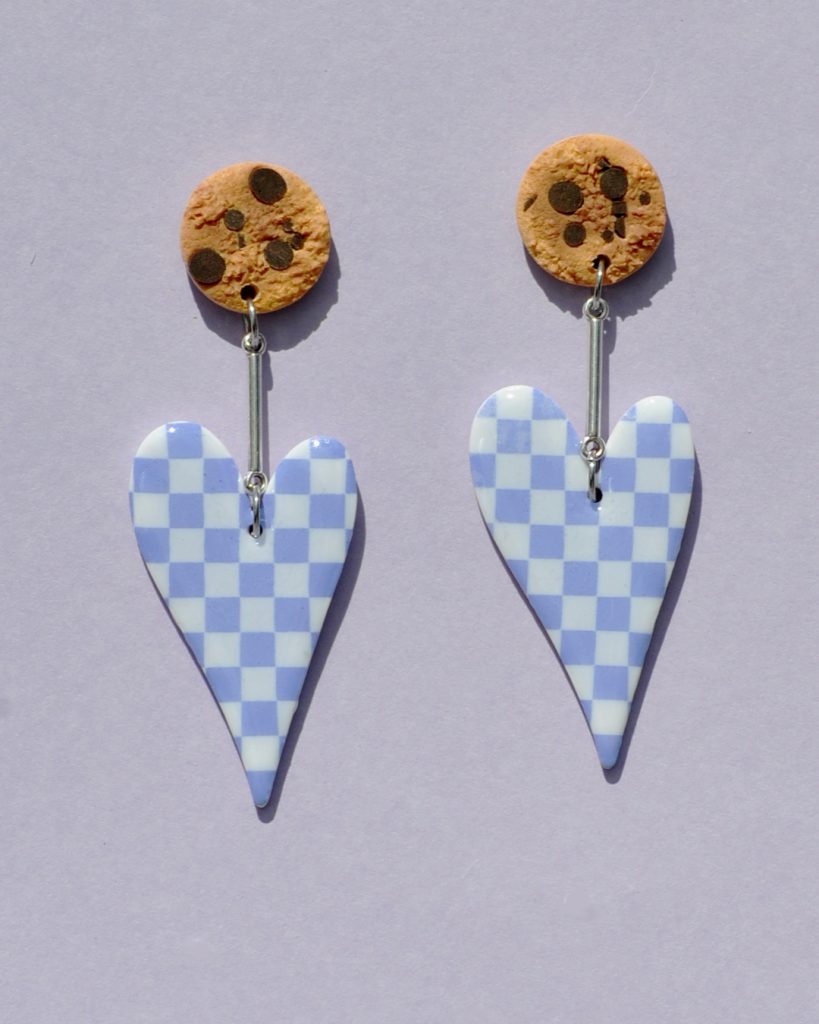 Paire de boucles d'oreilles en forme de cœur asymétrique allongé. Une puce cookie surmonte une tige en acier inoxydable argentée, surmontant la boucle au motif damier violet pâle sur fond blanc.