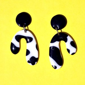 Paire de boucles d'oreilles en forme abstraite motif tâches de vache noires sur fond blanc, surmontée d'une puce ronde noire.
