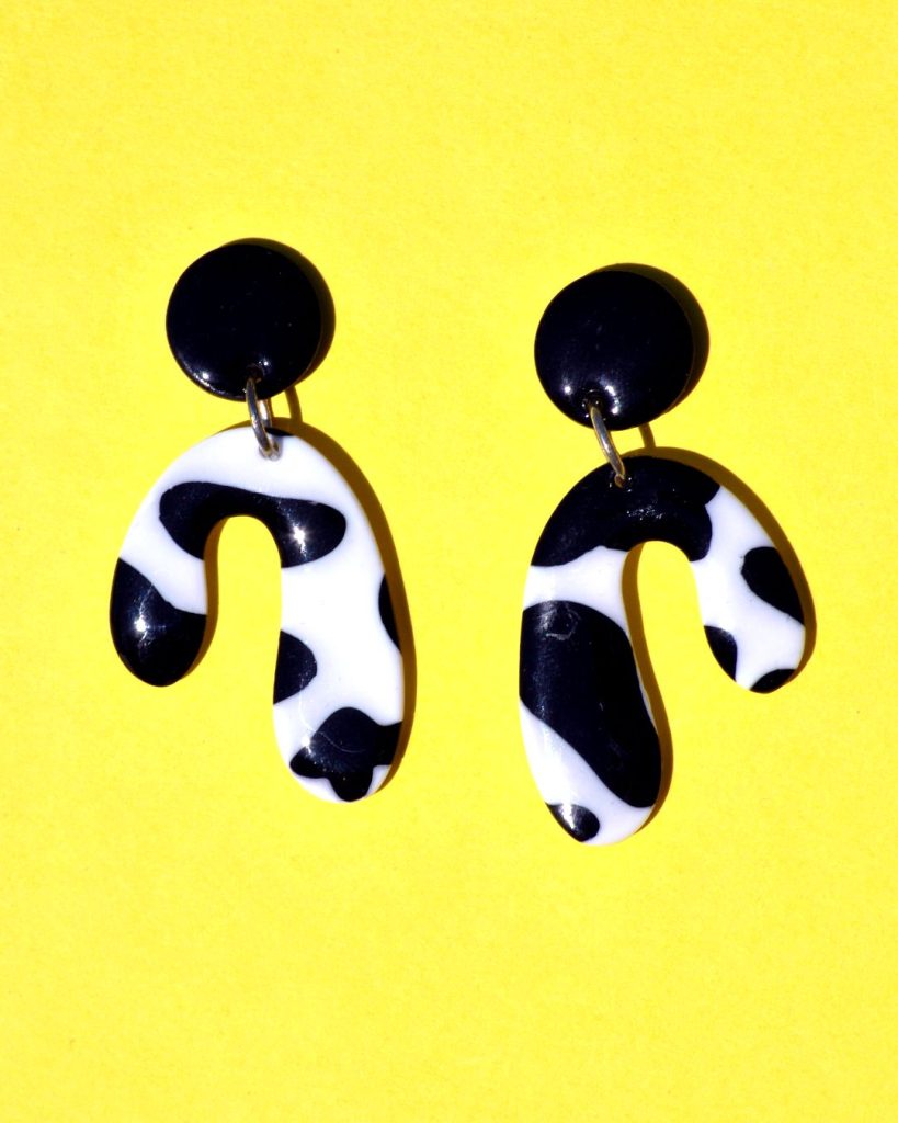Paire de boucles d'oreilles en forme abstraite motif tâches de vache noires sur fond blanc, surmontée d'une puce ronde noire.