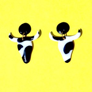 Boucles d'oreilles en forme de tête de taureau, avec tâches de vache noires sur fond blanc, surmontées de puces noires.