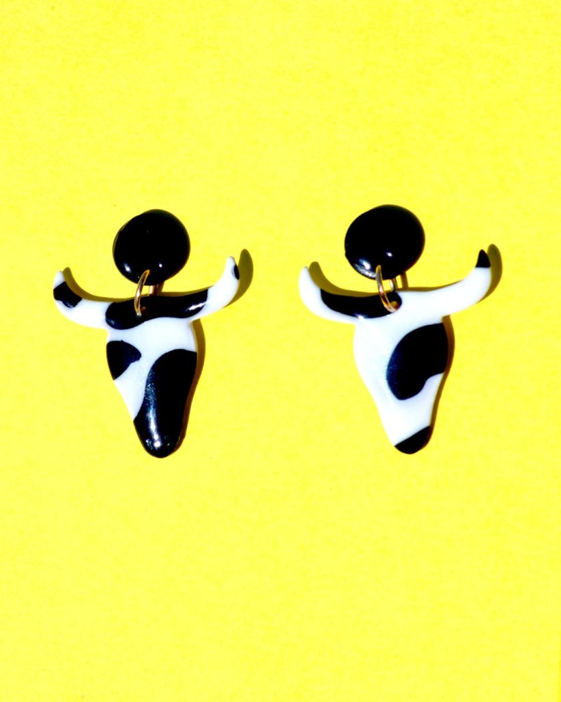 Boucles d'oreilles en forme de tête de taureau, avec tâches de vache noires sur fond blanc, surmontées de puces noires.