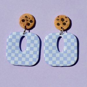 Paire de boucles d'oreilles en forme de carré rétro arrondi. La paire de boucles d'oreilles est ajourée au centre d'une ellipse. Une puce cookie surmonte une boucle motif damier blanc sur fond violet pâle.