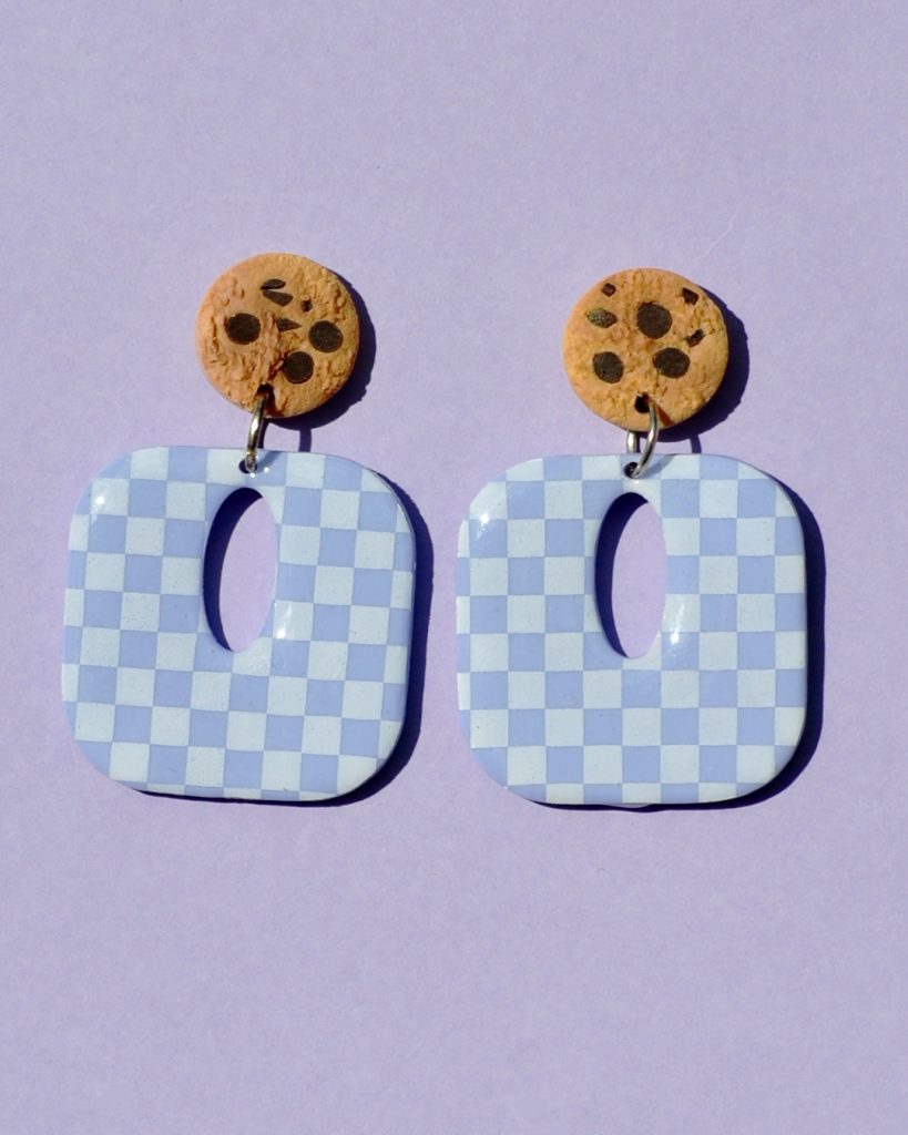 Paire de boucles d'oreilles en forme de carré rétro arrondi. La paire de boucles d'oreilles est ajourée au centre d'une ellipse. Une puce cookie surmonte une boucle motif damier blanc sur fond violet pâle.