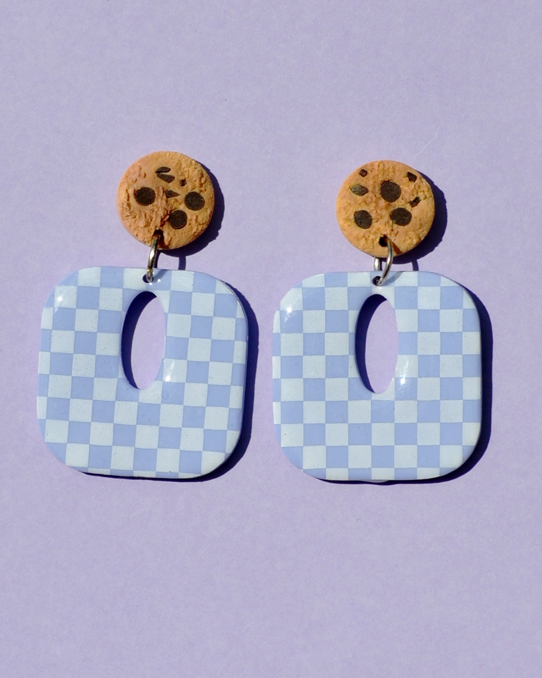 Paire de boucles d'oreilles en forme de carré rétro arrondi. La paire de boucles d'oreilles est ajourée au centre d'une ellipse. Une puce cookie surmonte une boucle motif damier blanc sur fond violet pâle.