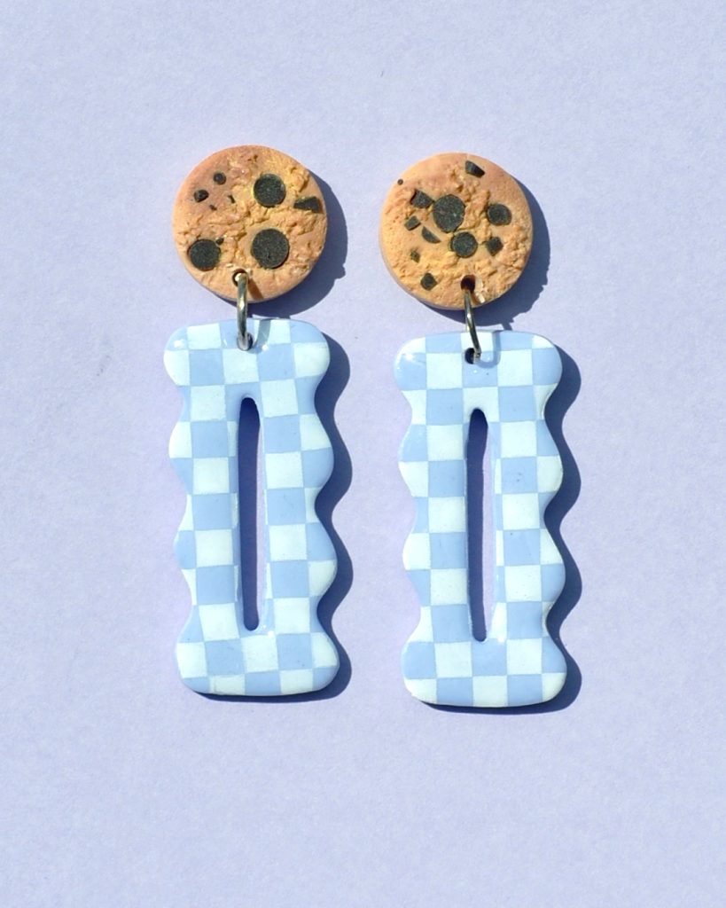 Paire de boucles d'oreilles en forme "petit lu" festonné allongé. La paire de boucles d'oreilles est ajourée au centre d'une ellipse très fine. Une puce cookie surmonte une boucle motif damier blanc sur fond violet pâle.