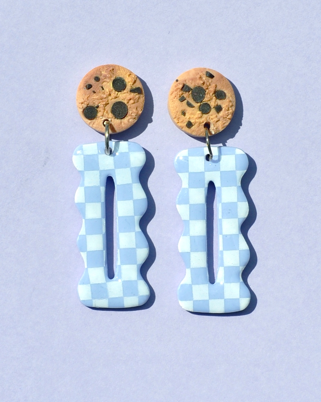 Paire de boucles d'oreilles en forme "petit lu" festonné allongé. La paire de boucles d'oreilles est ajourée au centre d'une ellipse très fine. Une puce cookie surmonte une boucle motif damier blanc sur fond violet pâle.