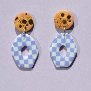 Paire de boucles d'oreilles en forme petit galet donut. La paire de boucles d'oreilles est ajourée au centre d'un cercle abstrait. Une puce cookie surmonte une boucle motif damier violet sur fond blanc.