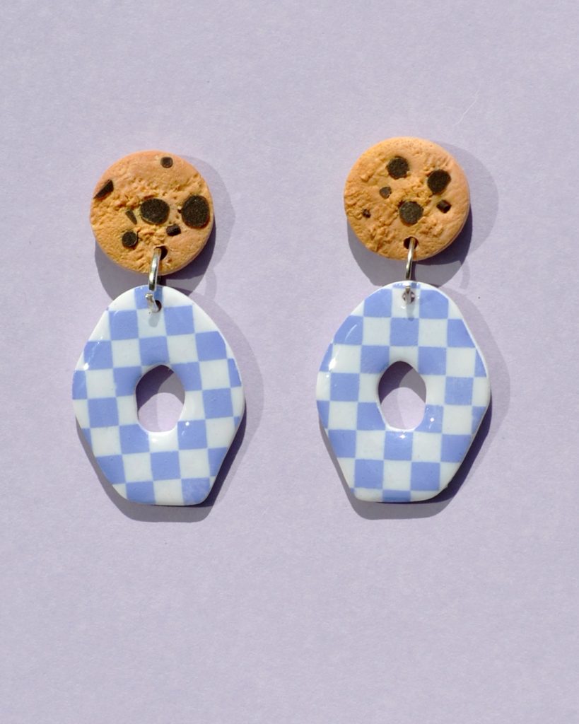 Paire de boucles d'oreilles en forme petit galet donut. La paire de boucles d'oreilles est ajourée au centre d'un cercle abstrait. Une puce cookie surmonte une boucle motif damier violet sur fond blanc.