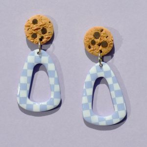 Paire de boucles d'oreilles en forme de triangle rétro déstructuré. La paire de boucles d'oreilles est ajourée au centre. Une puce cookie surmonte une boucle motif damier blanc sur fond violet pâle.