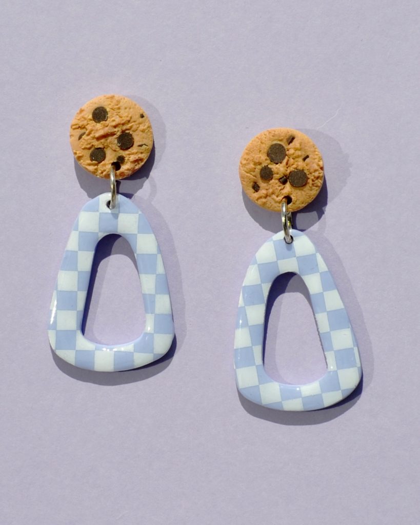 Paire de boucles d'oreilles en forme de triangle rétro déstructuré. La paire de boucles d'oreilles est ajourée au centre. Une puce cookie surmonte une boucle motif damier blanc sur fond violet pâle.