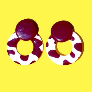 Paire de boucles d'oreilles en forme de cercle ajouré. Une grosse puce ronde en relief est collée sur le haut de la boucle. La boucle est au motif tâches de vache brunes sur fond blanc. La "puce" ronde est marron unie.